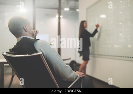 View from behind of a boardroom with a businessman in an office armchair closely observing how his female colleague in a defocused background Stock Photo