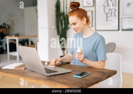 Happy young redhead woman is wiping laptop computer with sanitizer before starting work  Stock Photo