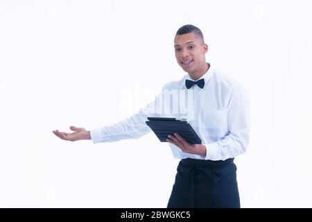 Young waiter doing a welcoming gesture while holding a tablet on a light background. Work and service concept. Stock Photo