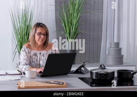 Focused mature woman using her laptop surrounded by a notebook, a tablet and a mug on a kitchen counter. Home office concept. Stock Photo