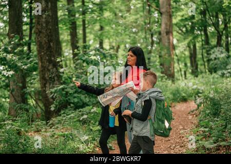 Beautiful mother with two kids are walking through forest, using a map and planning a hiking route. Happy family having fun together. Stock Photo