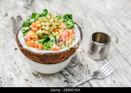 Salad with tomato, greens,  sprouted mung beans and coconut pulp served in half coconut on white wooden background