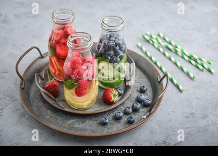 Infused water with fresh fruits, vegetables and berry in bottles on tray on gray background