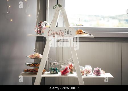 Rich offer of colorful candies in glass jars and on plates laying on white stand. Lettering Candy bar. Wedding or party concept. Stock Photo