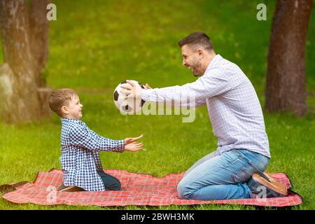 Fun picnic pastimes. Joyful dad and his cute little boy playing football on blanket in countryside Stock Photo