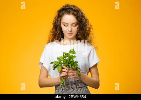 Closeup portrait of pretty caucasian girl with curly hair holding green fresh parsley isolated on colorful yellow background Stock Photo