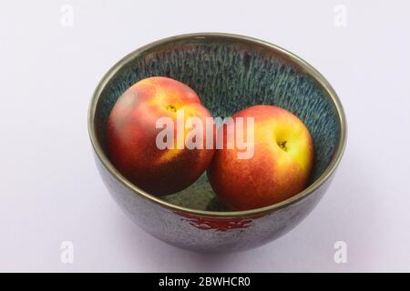 Two fresh raw nectarine fruits in blue bowl on lavender background Stock Photo