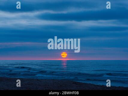 Dramatic sunset at Cavendish Beach, PEI National Park, Canada. Transient solar disk dazzles between dark storm clouds and azure ocean waters. Stock Photo