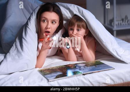 Woman and her little daughter reading book under blanket at night Stock Photo