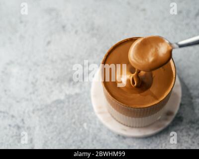 Trendy korean drink - dalgona coffee. Glass jar with cold or hot beverage - whipped instant coffee with milk poured from spoon. Gray cement background with copy space. Top view or flat lay. Stock Photo