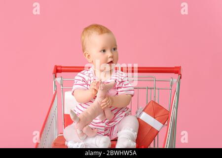Cute little baby with gifts sitting in shopping cart on color background Stock Photo