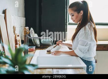 Young woman washes dishes with wooden brush with natural bristles at window in kitchen. Zero waste concept Stock Photo