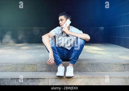 Young teenage boy chatting on his mobile phone outdoors sitting on city steps as he looks away to the side listening to the conversation with a though Stock Photo
