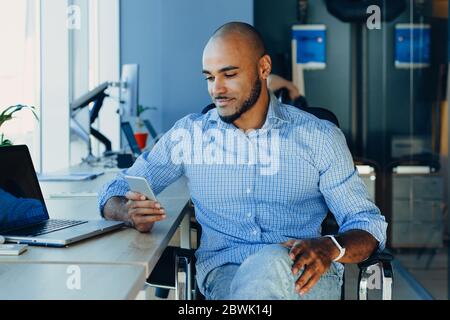 Handsome cheerful african american man in creative atmosphere using laptop sitting on a wooden table. Stock Photo
