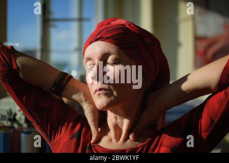 Female portrait of tired mature woman dressed in red clothes and sitting at the end of her work day on sewing machine and cotton bobbins background Stock Photo