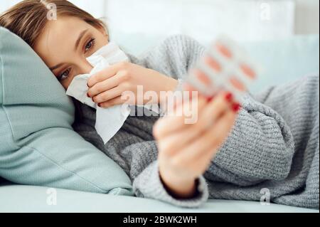 Sick Woman using paper tissue and holding pills. Flu. Sneezing into handkerchief. Headache. Virus .Medicines. Female Infected, blowing nose. Girl with Stock Photo