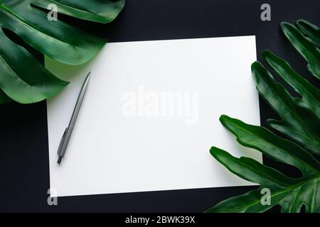 Blank white notepaper with tropical leaves laying on black table.Home office,workspace design backgrounds,flat lay,top view Stock Photo