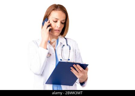 Portrait shot of confident female doctor talking on mobile phone and holding clipboard in her hand while standing at isolated white background. Stock Photo