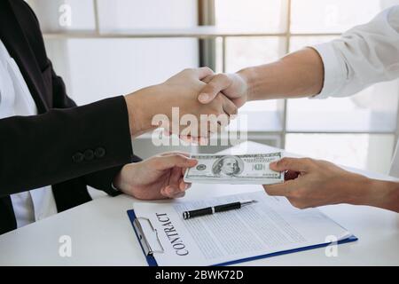 Two corporate businessmen shaking hands while one man places money on document in office room with corruption concept. Stock Photo