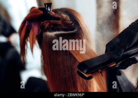 Hairdresser straightens redhead hair of woman with hair straightener tool in hair salon. Stock Photo