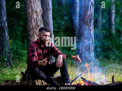 Man, hipster, hiker roast sausages on stick on bonfire in forest. Summer camping, hiking, vacation. Picnic, barbecue, cooking food concept. Guy with beard at picnic or barbecue. Stock Photo