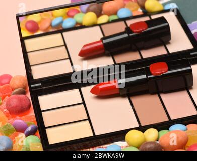 Make up concept. Beauty supplies, lipstick, beige palette, powder, rouge, concealer. Red, scarlet lipstick and palette with mirror lay on peachy background near dragee sweets. Stock Photo