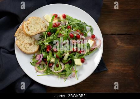 Arugula or rocket salad with pomegranate seeds, red onions and parmesan served with bread on a white plate on a dark rustic wooden table, copy space, Stock Photo