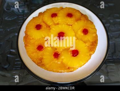 Golden yellow pineapple upside-down cake, a traditional sticky sponge cake with layers of sliced pineapple and red glace cherries, on a round plate Stock Photo