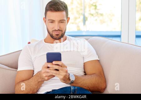 Handsome young man checking his mobile phone and reading messages while relaxing on sofa at home. Stock Photo