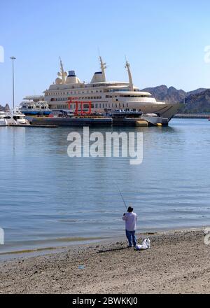 Local man fishing  on the beach with the Al Said royal yacht in the background,Muscat, Oman Stock Photo