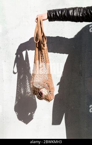 The delivery of food. Hand holding a mesh shopping bag with fresh food, hard shadows on the wall. Eco-friendly responsible lifestyle and shopping. A h Stock Photo