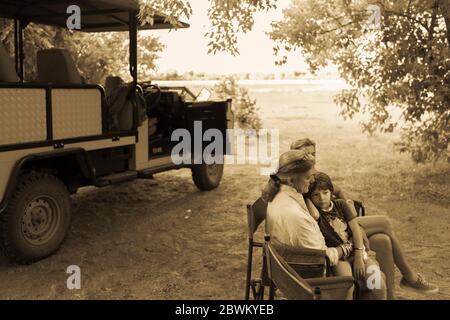 Three generations of a family seated in folding chairs by a safari jeep, under trees, monochrome. Stock Photo