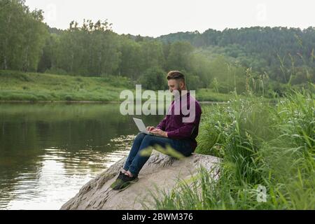 Freelance Concept, Remote Office. Young bearded man, businessman with laptop sits by river on summer day. Stock Photo
