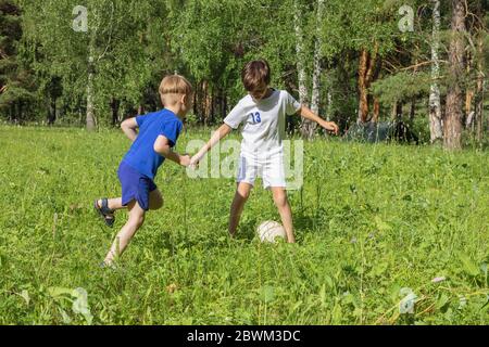 Two boys in uniform plays football on green meadow. Children run and kick soccer ball. Summer children outdoor games. Stock Photo