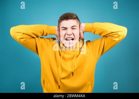 teenager in a yellow sweatshirt on a blue background pinches his ears with his hands and looks straight ahead Stock Photo
