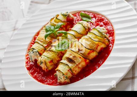 Stuffed zucchini roll-ups with soft ricotta cheese mixed with fresh baby spinach, basil served in tomato sauce on a white plate with fresh basil leaf