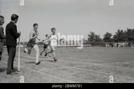 1950s, historical, summertime and a school sports day, teenage boys competing in a sprint race, with a fellow pupil holding the finishing tape as the winning runner nears the line. Stock Photo