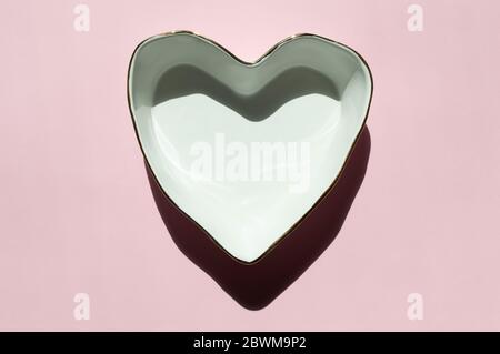 White ceramic clean empty plate with golden boarders in the shape of heart on a light pink background with sunny shadows. Geometrical shape. Pastel Stock Photo