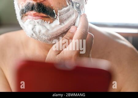Portrait of a busy and distracted young man shaving his beard while texting on his smartphone. Man uses smartphone as mirror when he shaves. Stock Photo