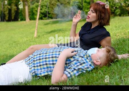 Young boy with surgical mask lying on the grass in the park near a sitting girl who smokes. Child dressed in a plaid shirt lying on the green field ne Stock Photo