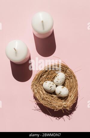 Two new white candles with sunny shadows near three eggs with tiny golden spots inside brown nest on the light pink background. Creative trendy Stock Photo