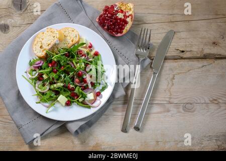 Arugula or rocket salad with pomegranate seeds, red onions and parmesan served on a white plate with cutlery on a rustic wooden table, copy space, hig Stock Photo