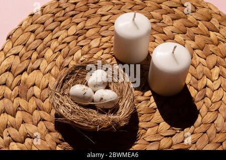 Creative composition of two white candles and three tiny eggs decorated with golden spots inside nest on the light brown mat, handmade from water Stock Photo