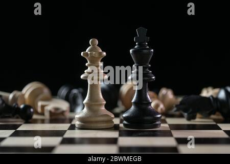 Chess pieces queen and king in front of a of fallen chessmen after a battlefield on a chessboard against a black background, concept of abuse of power Stock Photo