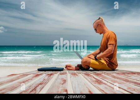 Young guy with long hair by the sea on a sunny day, working using his laptop, working remotely with a business. Bloner, freelancer, work through the I Stock Photo