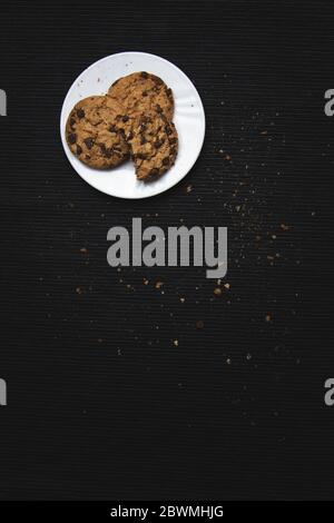Chocolate chip cookies in a white plate, on black fabric background full of crumbles and chocolate chips. Unhealthy food, delicious treat, high calori Stock Photo