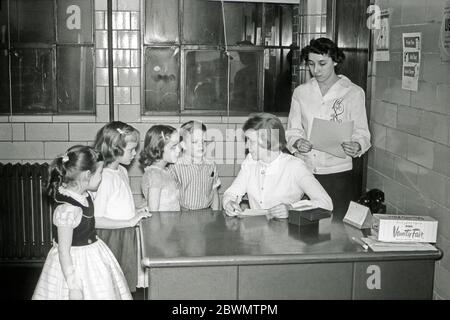 Education in the USA in the 1950s – here in a tiled office a nurse or health visitor is seeing and questioning four children in elementary school. She is using a card index system. On her desk is a swab in a glass tube and a box of paper tissues. There are health related posters on the wall. Stock Photo