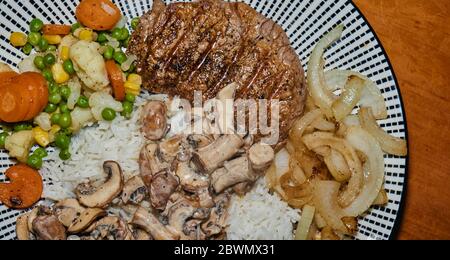 Complete meal of a grilled pork steak with roasted onions, vegetable mix of peas, carrots, corn and cauliflower, on rice with mushrooms in cream sauce Stock Photo