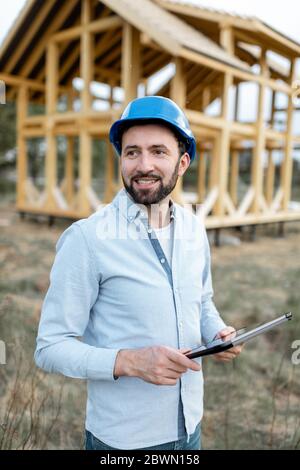 Portrait of an architect or builder with digital touchpad in front of the wooden house structure. Building and designing wooden frame house