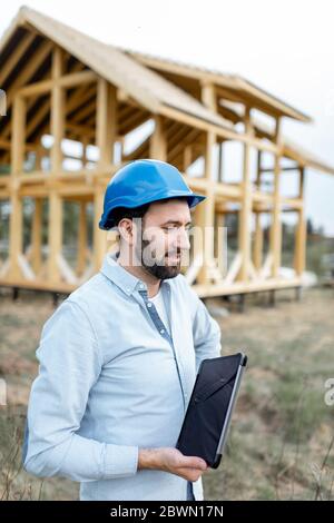 Portrait of an architect or builder with digital touchpad in front of the wooden house structure. Building and designing wooden frame house Stock Photo